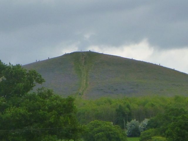 Steep green hill under cloudy sky with trees below.