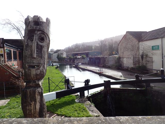 Wooden sculpture beside canal lock and rural buildings.